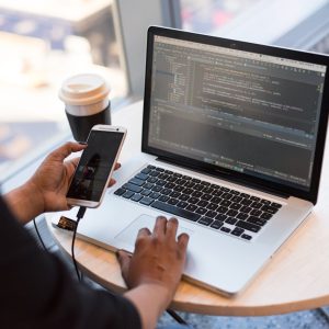 Developer working remotely, coding on a laptop with phone in hand, showcasing modern work culture.
